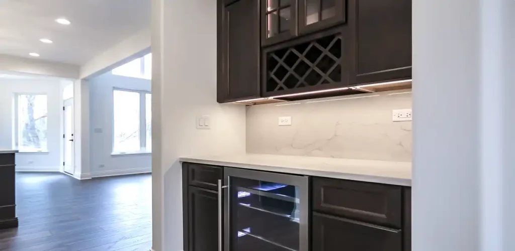 A completed kitchen wet bar in a Bethesda home renovation featuring dark wood custom cabinetry, integrated wine storage, under-cabinet LED lighting, and a stainless steel beverage refrigerator.
